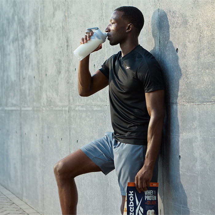 A man in athletic wear leans against a concrete wall, sipping Reebok ReeBuild Whey Protein + Collagen and holding its branded pack, looking relaxed after his workout.