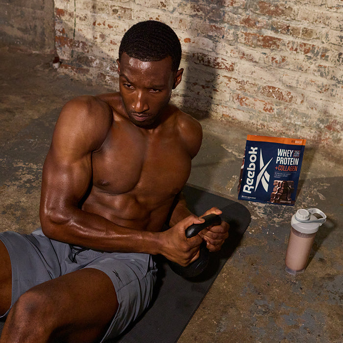 A muscular man in gray shorts sits on a black exercise mat by a brick wall, holding a kettlebell. Beside him are Reebok ReeBuild Whey Protein + Collagen and a shaker bottle, highlighting post-workout muscle recovery.