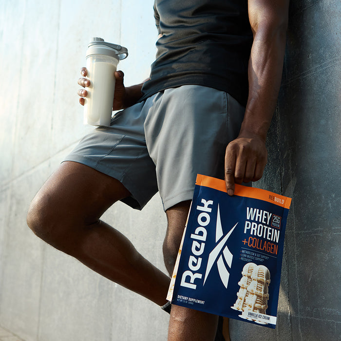Wearing gray shorts and a black shirt, a person leans against a wall holding a shaker of milk and a Reebok ReeBuild Whey Protein + Collagen pouch, ready to support muscle recovery.