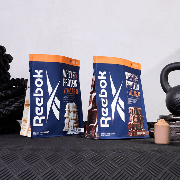 Two bags of Reebok ReeBuild Whey Protein + Collagen, featuring collagen peptides for muscle recovery, are shown on a black gym mat with a kettlebell, rope, and scoop—available in vanilla ice cream and chocolate brownie flavors.