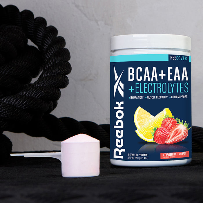 A container of Reebok ReeCover BCAA + EAA + Electrolytes in strawberry lemonade flavor sits by a white scoop of powder, with black exercise ropes behind—ideal for muscle recovery and loaded with essential amino acids.