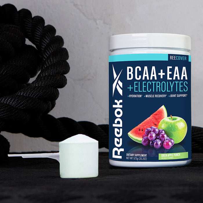 A container of Reebok ReeCover BCAA + EAA + Electrolytes in green apple punch flavor sits on a dark surface next to a scoop, showcasing its balanced blend of BCAAs and Essential Amino Acids for optimal workout recovery.