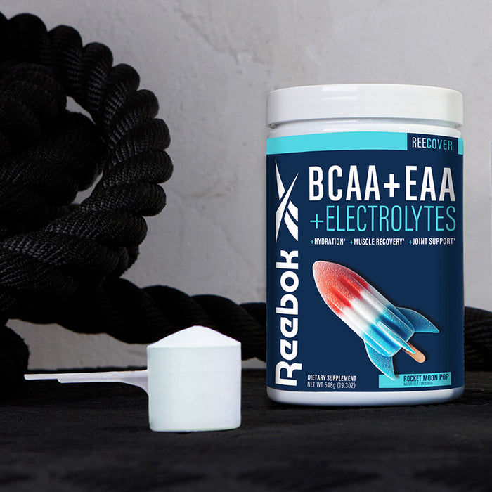 A tub of Reebok ReeCover BCAA + EAA + Electrolytes powder, featuring Essential Amino Acids for muscle recovery, is displayed next to a rocket popsicle image, a scoop of powder, and a thick black rope in the background.