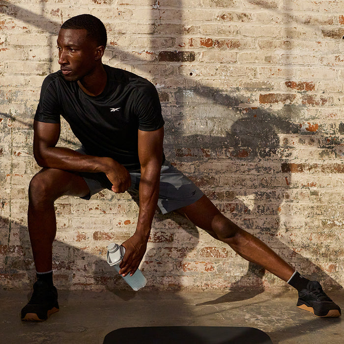 A man in a black t-shirt and gray shorts does a side lunge stretch against a sunlit brick wall, holding Reebok ReeCover BCAA + EAA + Electrolytes—perfect for muscle recovery after a tough workout. Shadows fall on the wall behind him.