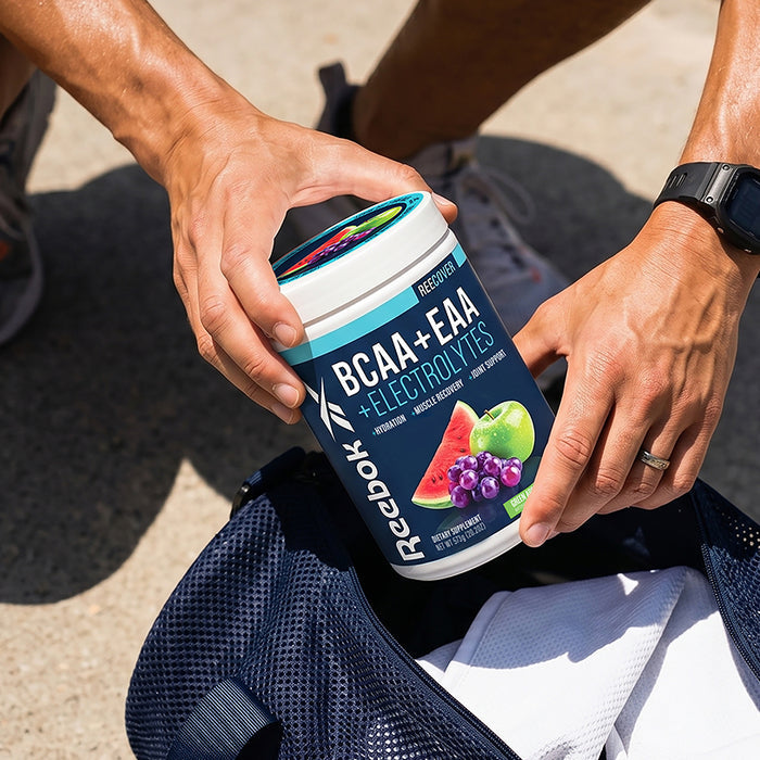 A person packs a navy blue gym bag with Reebok ReeCover BCAA + EAA + Electrolytes, featuring images of watermelon, grape, and apple. They wear a smartwatch and wedding ring, ready for workout recovery.
