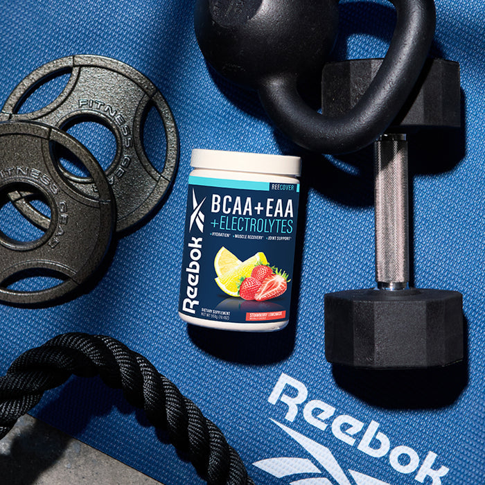 A container of Reebok ReeCover BCAA + EAA + Electrolytes rests on a blue Reebok mat, surrounded by a dumbbell, weight plates, and a black workout rope—your go-to partner for muscle recovery.