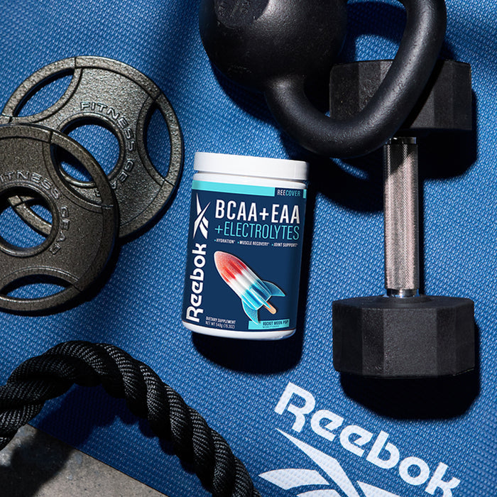 A container of Reebok ReeCover BCAA + EAA + Electrolytes, a supplement for muscle recovery, rests on a blue Reebok exercise mat next to a black dumbbell, kettlebell, gym rope, and weight plates.