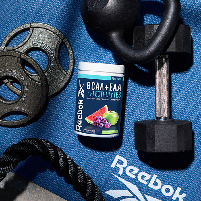 A tub of Reebok ReeCover BCAA + EAA + Electrolytes for workout recovery rests on a blue Reebok exercise mat, surrounded by a dumbbell, kettlebell, weight plates, and a black exercise rope.