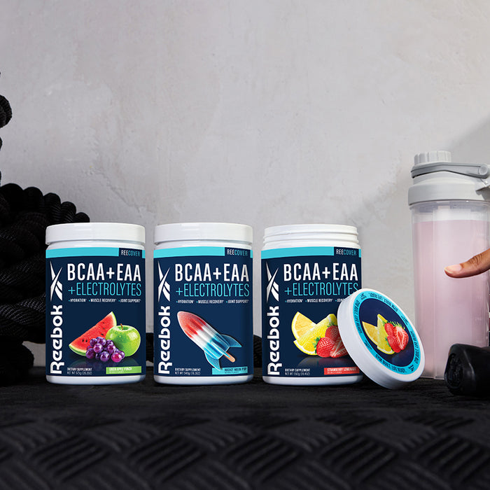 Three Reebok ReeCover BCAA + EAA + Electrolytes tubs in assorted flavors are shown on a black surface beside a shaker bottle and a hand holding its lid. These supplements support muscle recovery with essential amino acids. Ropes appear in the background.