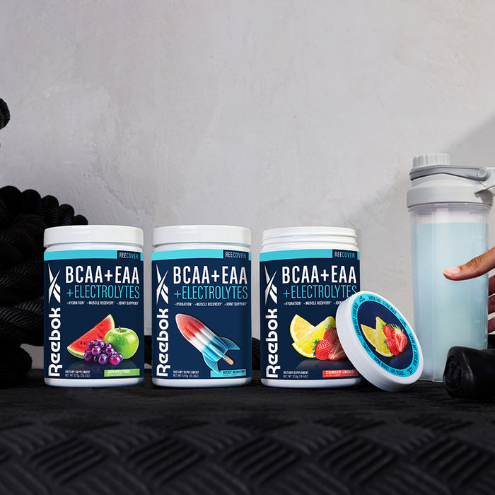 Three tubs of Reebok ReeCover BCAA + EAA + Electrolytes, featuring fruit designs, sit on a counter next to a shaker bottle and black jump rope, emphasizing muscle recovery support. A hand reaches for one tub's open lid.