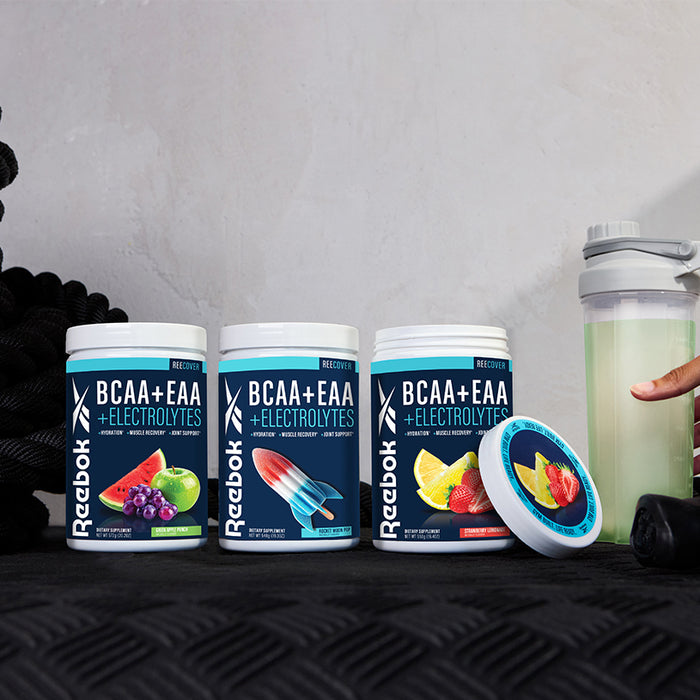 Three Reebok ReeCover BCAA + EAA + Electrolytes containers in grape, popsicle, and watermelon flavors are shown on a table with a shaker bottle and coiled black rope—ideal for workout recovery.