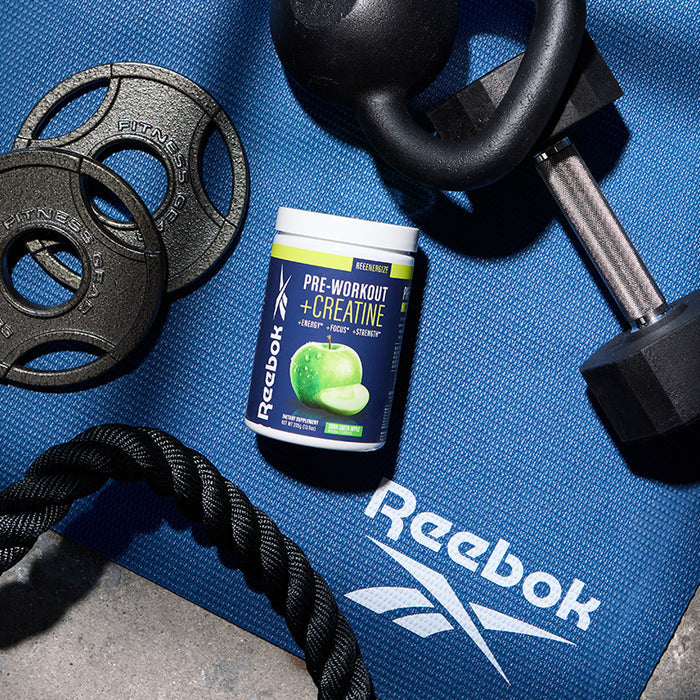 A container of Reebok ReeEnergize Pre-Workout + Creatine rests on a blue Reebok mat, surrounded by a kettlebell, dumbbell, weight plates, and battle rope. Clean caffeine delivers an energizing boost for your workout.