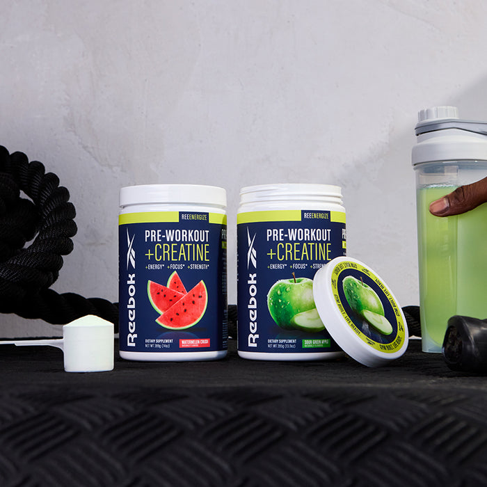 Two Reebok ReeEnergize Pre-Workout + Creatine tubs—watermelon and green apple—sit on a tire, with a white scoop, shaker bottle of green drink, ropes, and a hand capturing this energizing Reebok workout setup.