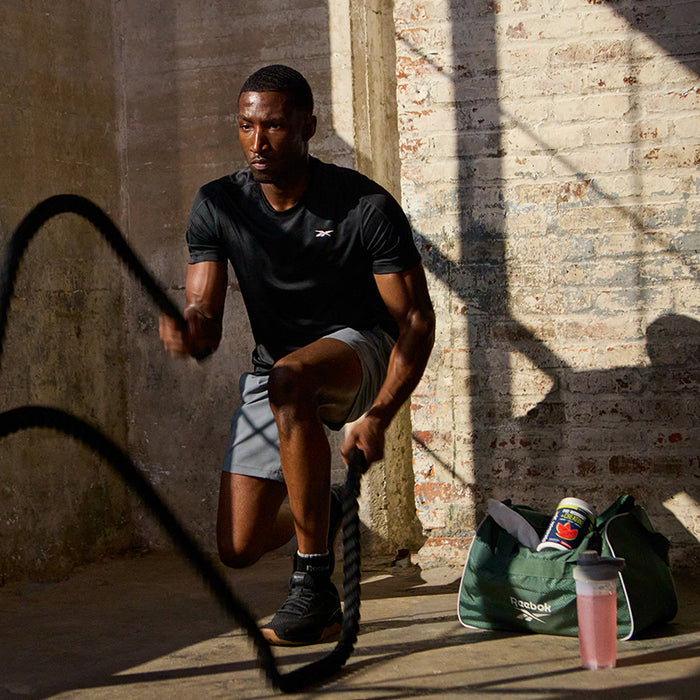 A man in athletic wear does battle rope exercises in a sunlit, industrial-style gym; on the floor are a green Reebok gym bag and a pink shaker with Reebok ReeEnergize Pre-Workout + Creatine.