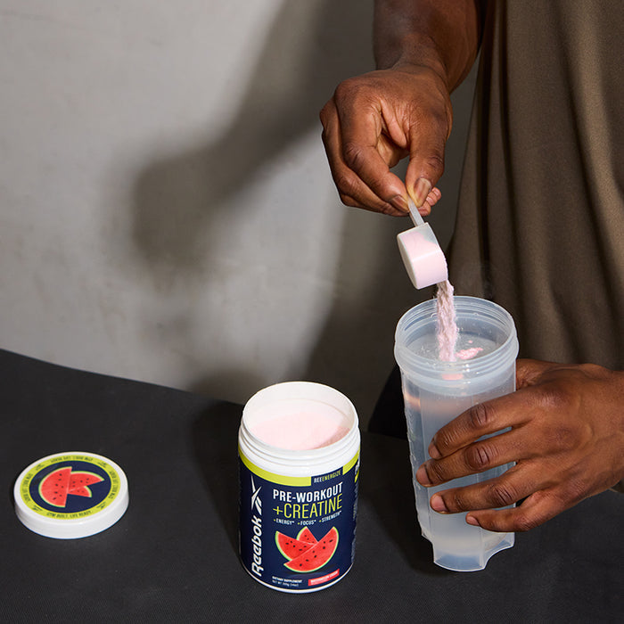 A person scoops pink Reebok ReeEnergize Pre-Workout + Creatine powder from a Reebok container with a watermelon lid and pours it into a clear shaker bottle.