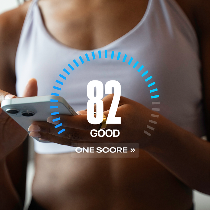 A woman in a white sports bra uses a Reebok Smart Ring to monitor her biometrics. Her smartphone displays an 82 score labeled GOOD and ONE SCORE below a blue semi-circular bar, showcasing the Reebok wearable fitness tracker.