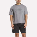 A man wearing a loose Reebok x F45 Training Graphic T-Shirt in gray and black shorts stands facing forward against a plain white background.