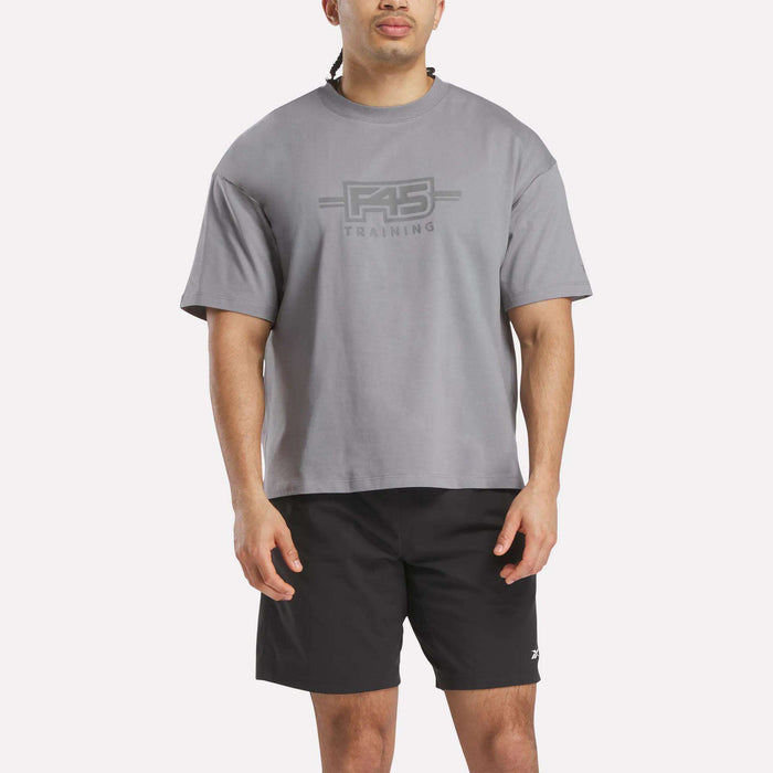 A man wearing a loose Reebok x F45 Training Graphic T-Shirt in gray and black shorts stands facing forward against a plain white background.