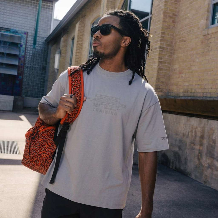 A man with long dreadlocks, sunglasses, and black shorts wears a Reebok x F45 Training Tonal Graphic T-Shirt and carries a red patterned backpack over his shoulder while walking outside near a brick building.