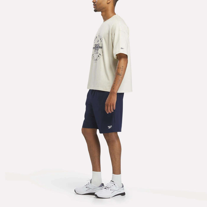A person stands sideways wearing a Reebok X F45 Training Graphic T-Shirt in beige, paired with navy shorts, white socks, and white sneakers. The background is plain white.