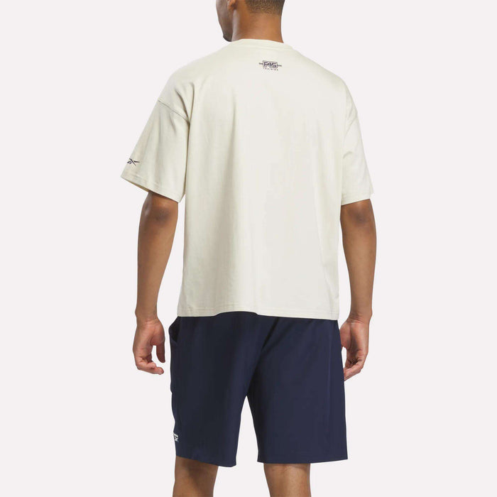 A person stands facing away, showcasing the back of the outfit against a plain light background, wearing a loose-fitting Reebok X F45 Training Graphic T-Shirt in white paired with navy blue shorts.