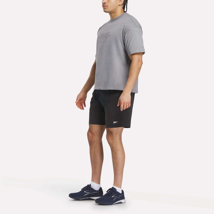 A man stands wearing a Reebok x F45 Training Graphic T-Shirt in gray, paired with black shorts and navy blue Reebok athletic shoes, against a plain light background.