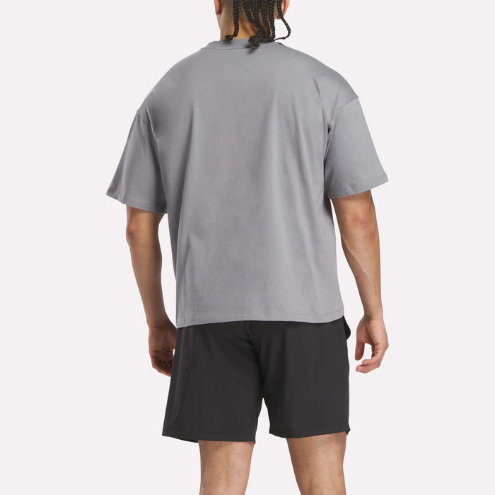 A person with braided hair is seen from behind, wearing a loose-fitting Reebok x F45 Training Tonal Graphic T-Shirt and black shorts, standing against a plain light background.