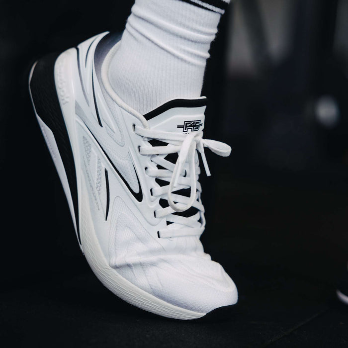 A close-up of a person wearing the white Reebok x F45 Training Nano X5 Edge with black accents and white laces, paired with white socks. The modern sneaker displays sleek design details and branding on the tongue.