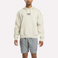 A person wearing the Reebok x F45 Training Relaxed Fleece Hoodie in Linen—a cream-colored, relaxed-fit hoodie featuring "F45 Training" on the chest—paired with light gray athletic shorts, stands against a plain white background.