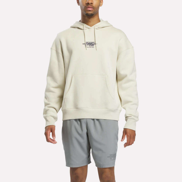 A person wearing the Reebok x F45 Training Relaxed Fleece Hoodie in Linen—a cream-colored, relaxed-fit hoodie featuring "F45 Training" on the chest—paired with light gray athletic shorts, stands against a plain white background.