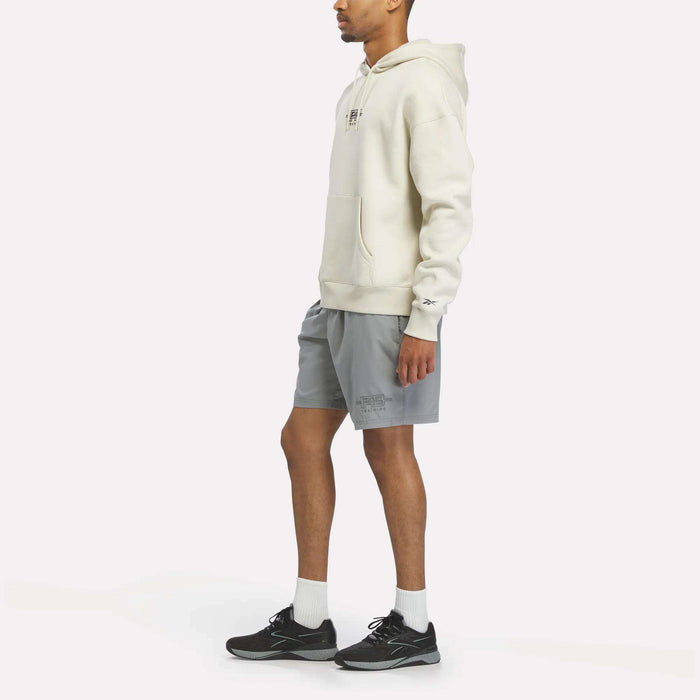 Wearing a Reebok x F45 Training Relaxed Fleece Hoodie in cream, gray shorts, white socks, and black sneakers, a person stands facing sideways against a plain white background.