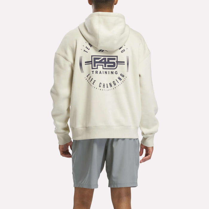 A person wearing a Reebok x F45 Training Relaxed Fleece Hoodie in cream, featuring "F45 Training" and "Life Changing" text on the back, pairs it with light gray shorts while standing with their back to the camera. The Reebok hoodie offers a roomy fit and ultimate comfort thanks to its relaxed fleece design.