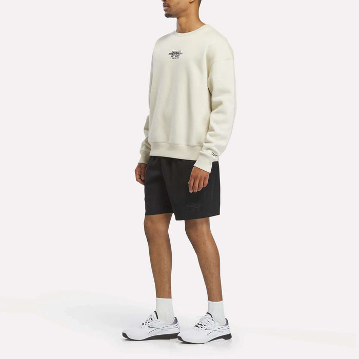A person wearing a cream Reebok x F45 Training Relaxed Graphic Sweatshirt, black shorts, white socks, and white sneakers stands against a plain white background. They are facing slightly to the right with their arms relaxed at their sides.