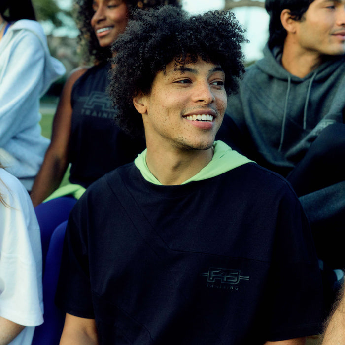 A young man with curly hair and freckles smiles outdoors with friends, all casually dressed in Reebok x F45 Training Relaxed T-Shirts from Reebok.