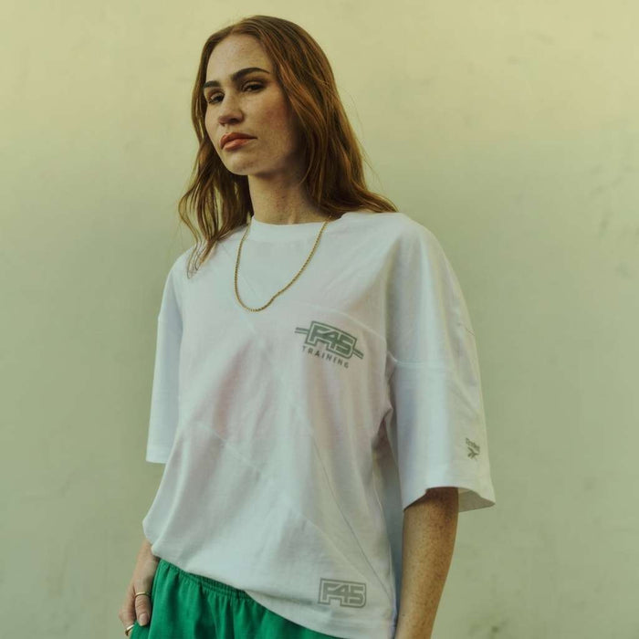 A woman with long brown hair wears the Reebok x F45 Training Relaxed T-Shirt and green shorts, standing against a plain light background and embodying the laid-back style of Reebok’s collaboration with F45 Training.