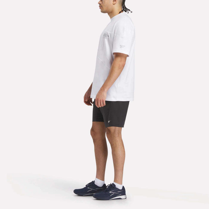 A person stands sideways against a plain, light background, wearing the Reebok x F45 Training Relaxed T-Shirt, black shorts, and navy blue Reebok performance athletic shoes.