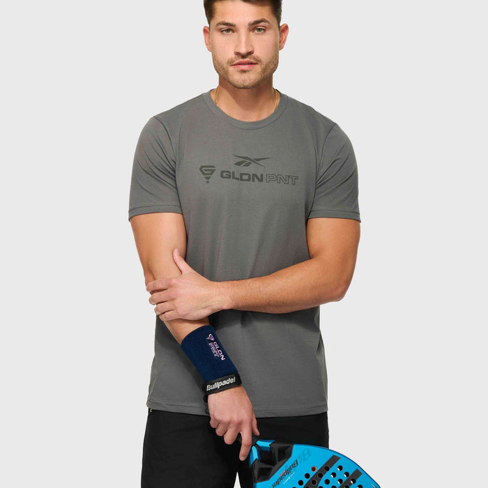 A man wears a Reebok x GLDN PNT Jersey Tee and black shorts, holding a blue padel racket in his left hand. He also has a black wristband on his right wrist and stands against a plain light background.