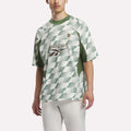 A person stands against a plain background wearing the Reebok x Manors Men’s Mockneck Polo—a green and white checkered short-sleeve jersey crafted from performance stretch fabric with a MANORS chest logo—paired with white pants.