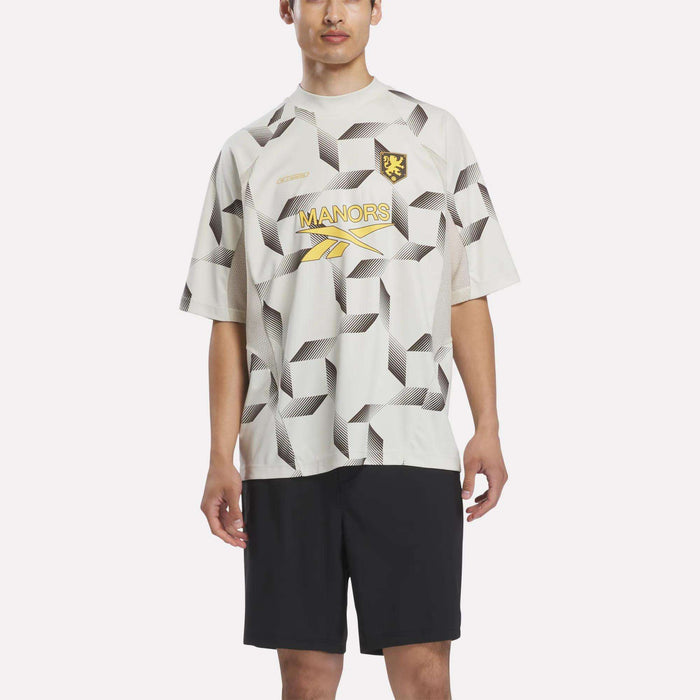 A person stands wearing the Reebok x Manors Men’s Mockneck Polo, featuring a geometric white and gray pattern, a black lion crest, and “MANORS” branding. The polo is crafted from performance stretch fabric and paired with black shorts. The background is plain white.