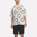 A person stands wearing the Reebok x Manors Men’s Mockneck Polo, featuring a geometric white and gray pattern, a black lion crest, and “MANORS” branding. The polo is crafted from performance stretch fabric and paired with black shorts. The background is plain white.