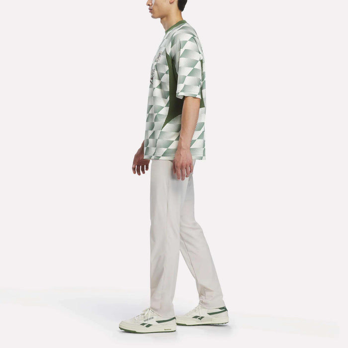 A person standing sideways, wearing the Reebok x Manors Men’s Mockneck Polo by Reebok—featuring a geometric-patterned green and white design crafted from performance stretch fabric—paired with light pants and white sneakers with green accents, set against a plain white background.