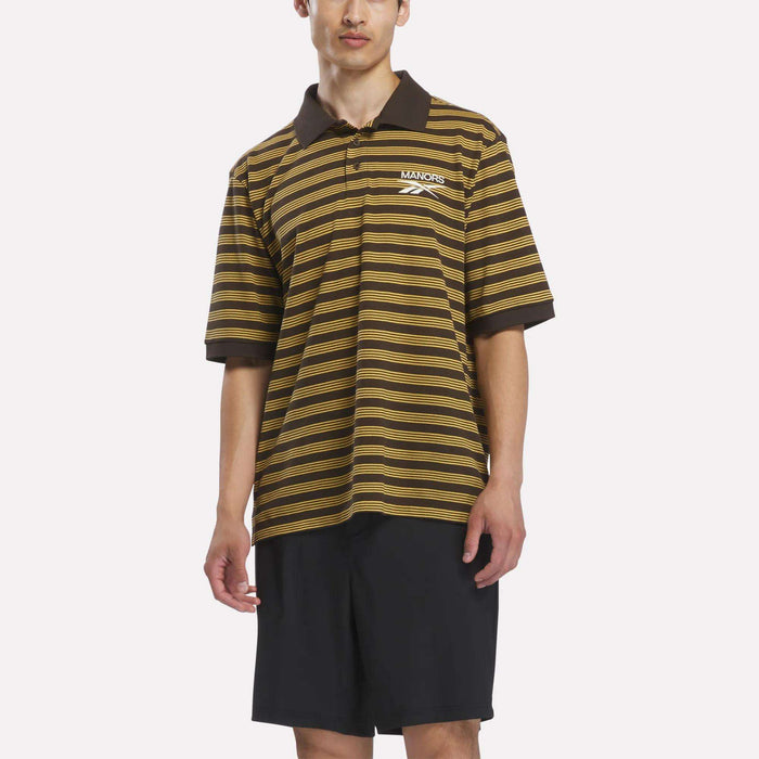 A person wearing the Reebok x Manors Men’s Pique Polo in Espresso/Gorse and black shorts stands against a plain white background.