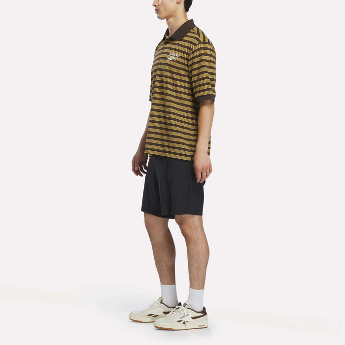 A person wearing a Reebok x Manors Men’s Pique Polo in brown and black stripes, paired with black shorts, white socks, and white sneakers stands against a plain white background.