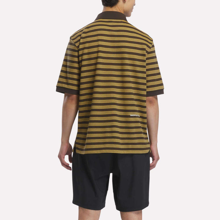 A person stands facing away, wearing a brown and yellow striped Reebok x Manors Men’s Pique Polo and black shorts against a plain light background.