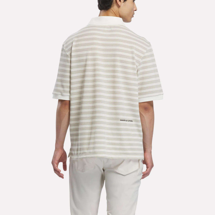 A man is shown from the back, standing against a plain white background, wearing the Reebok x Manors Men’s Pique Polo—a light-colored, short-sleeve striped golf shirt—paired with white pants. This classic look showcases the modern style of Reebok’s men’s golf polo, perfect for both on and off the course.