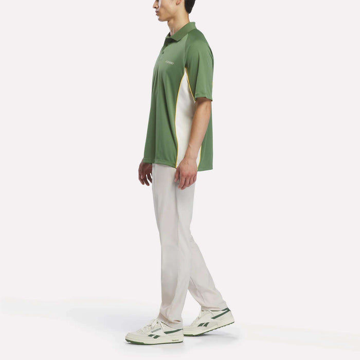 A man stands sideways against a plain white background, wearing the Reebok x Manors Men’s Polo in green with white accents, paired with light-colored pants and white sneakers featuring green details.