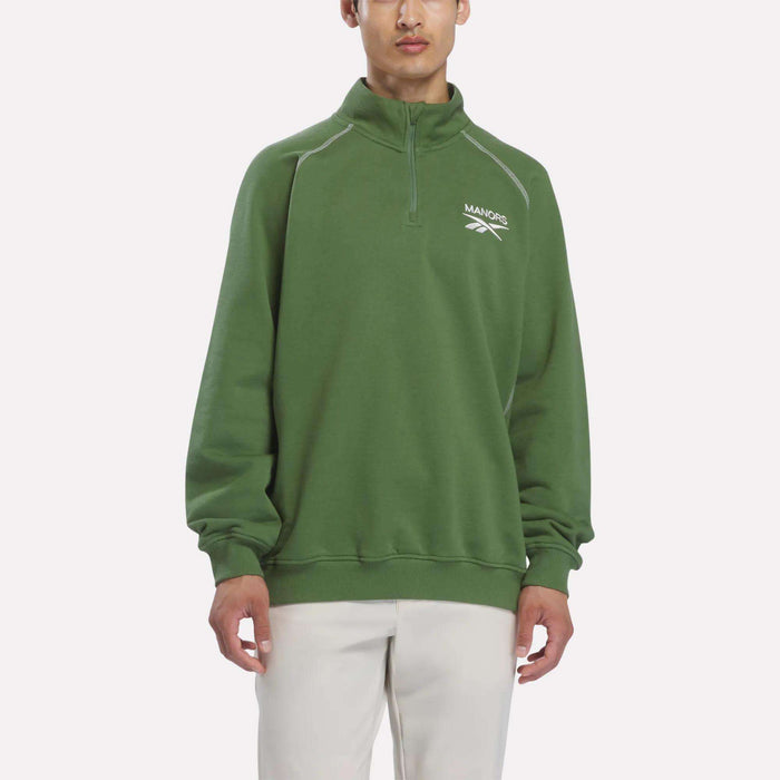 A person wearing the Reebok x Manors Men’s Quarter-Zip Pullover in green, with a white MANORS logo, paired with white pants, stands against a plain light background.