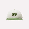The Reebok x Manors Men’s Retro Crown Cap features a crisp white design with a green brim and green embroidered MANORS text and logo on the front. This classic golf cap from Reebok includes an adjustable snapback for a perfect fit and is shown against a simple light background.