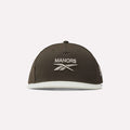 Introducing the Reebok x Manors Men’s Retro Crown Cap, a dark brown baseball cap featuring MANORS and the Reebok logo embroidered in white on the front. This cap showcases a retro crown silhouette, complemented by a light-colored brim, an adjustable snapback closure, and a small logo on the left side.
