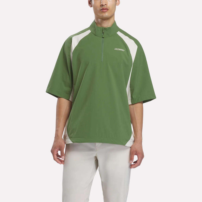 A man wearing the Reebok x Manors Men’s Tour Shirt by Reebok in green and white short-sleeves, paired with light-colored pants, stands against a plain white background.